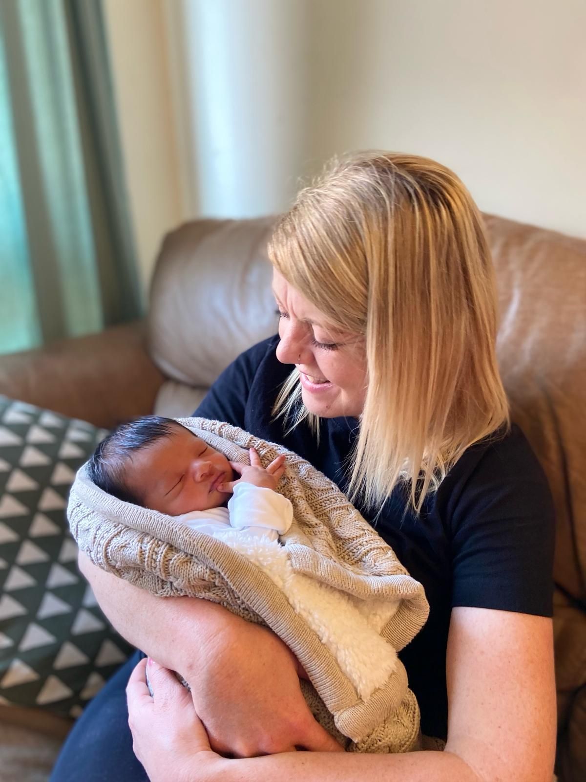 Woman holding a swaddled baby; tan blanket, brown couch, light blond hair, smiling.