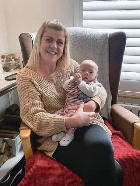 Woman holding a baby in a chair. Both smiling, the woman wears a tan sweater, the baby has on a patterned outfit.