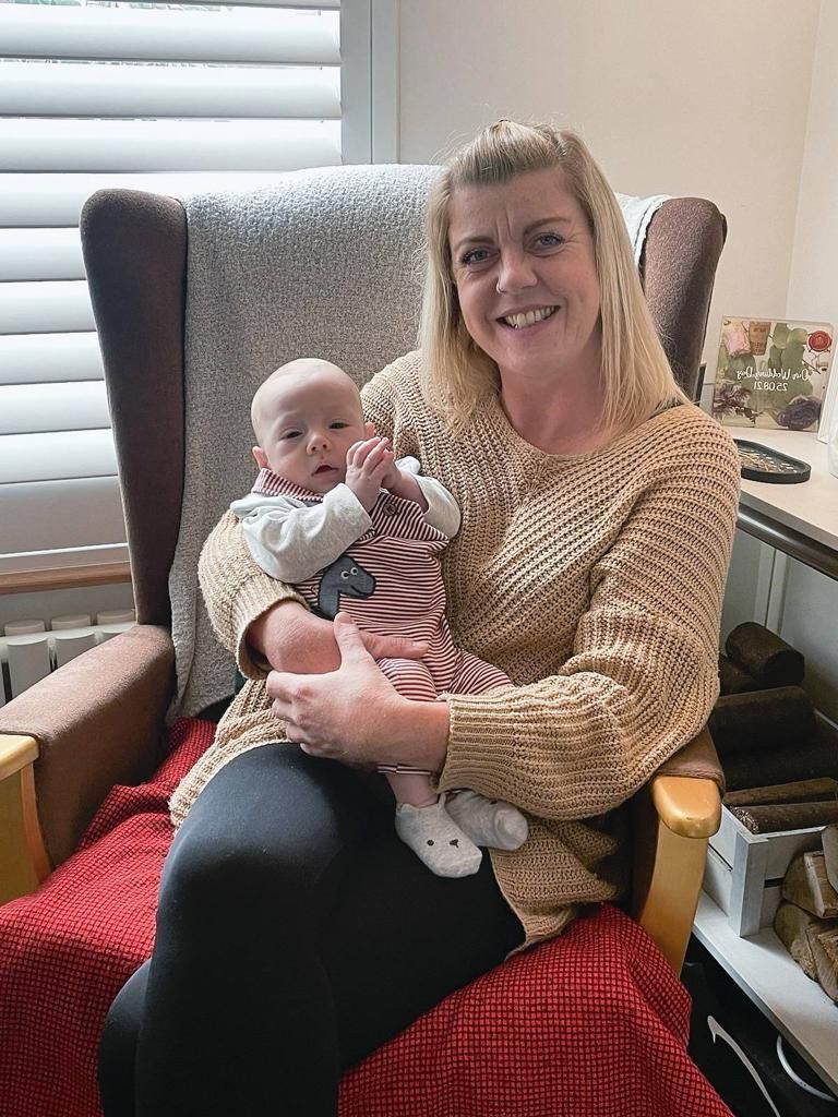 Woman sitting with a baby in a chair, both smiling. The woman wears a tan sweater, baby wears a striped outfit.