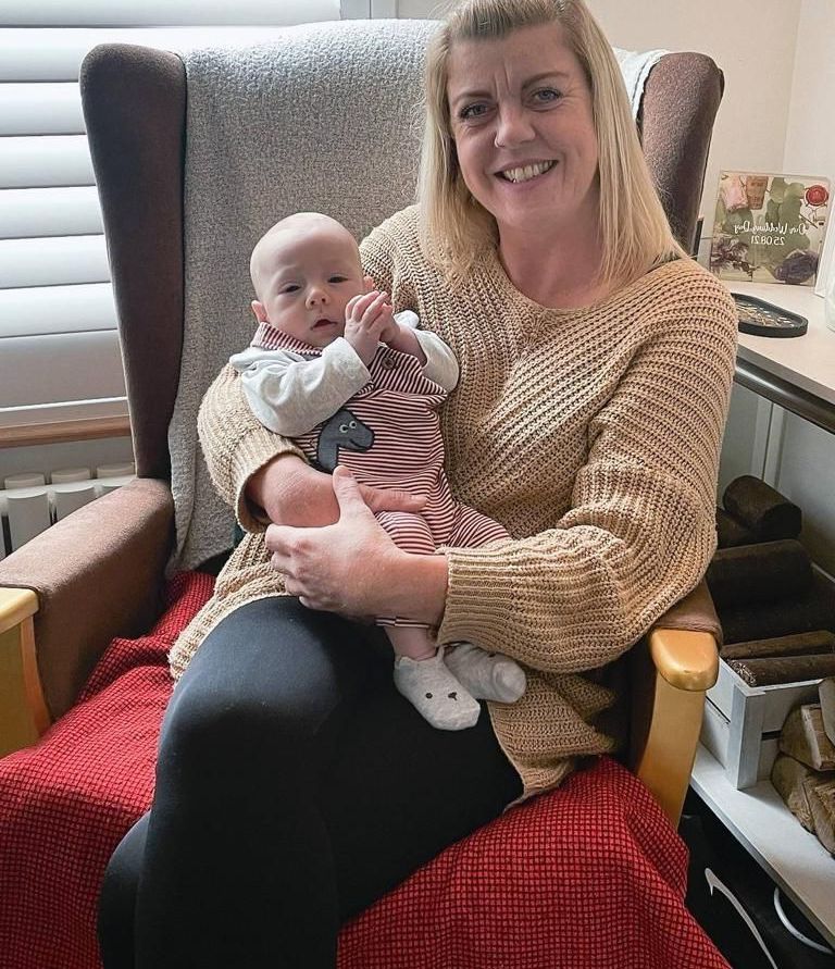 Woman in beige sweater holds a baby, both smiling, seated in a wooden armchair with red blanket.