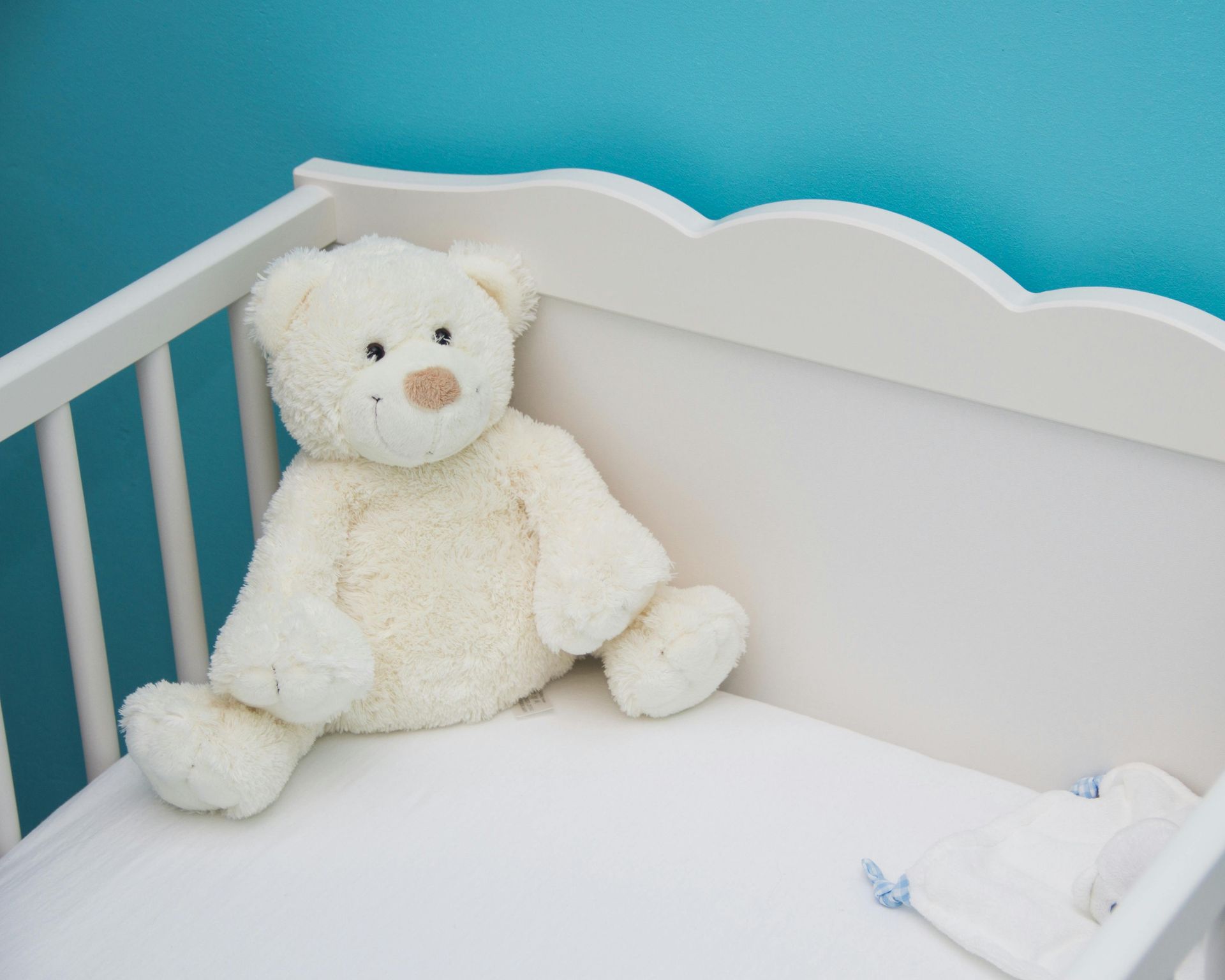 White teddy bear in a white crib with a blue wall in the background.