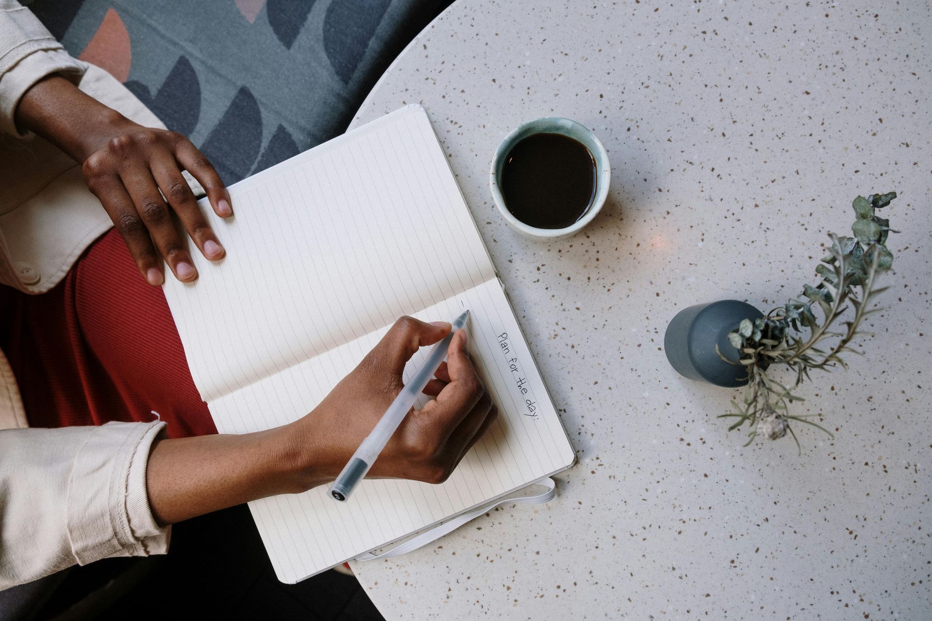 Person writing in a notebook, pen in hand, coffee cup and plant on the table.
