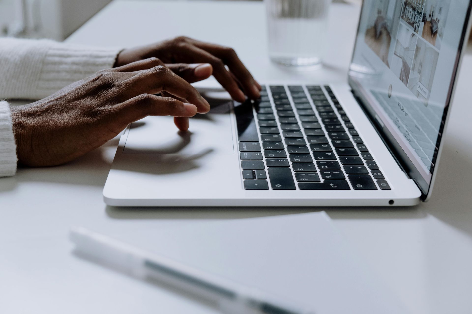 Person typing on a laptop with hands visible, white table, and pen.