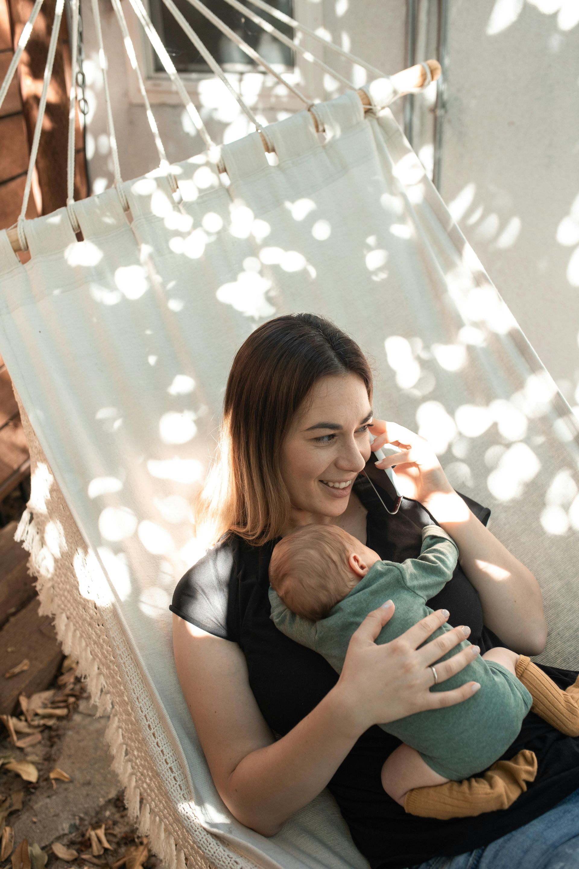 Woman holding baby in hammock, talking on phone, sunny outdoor setting.