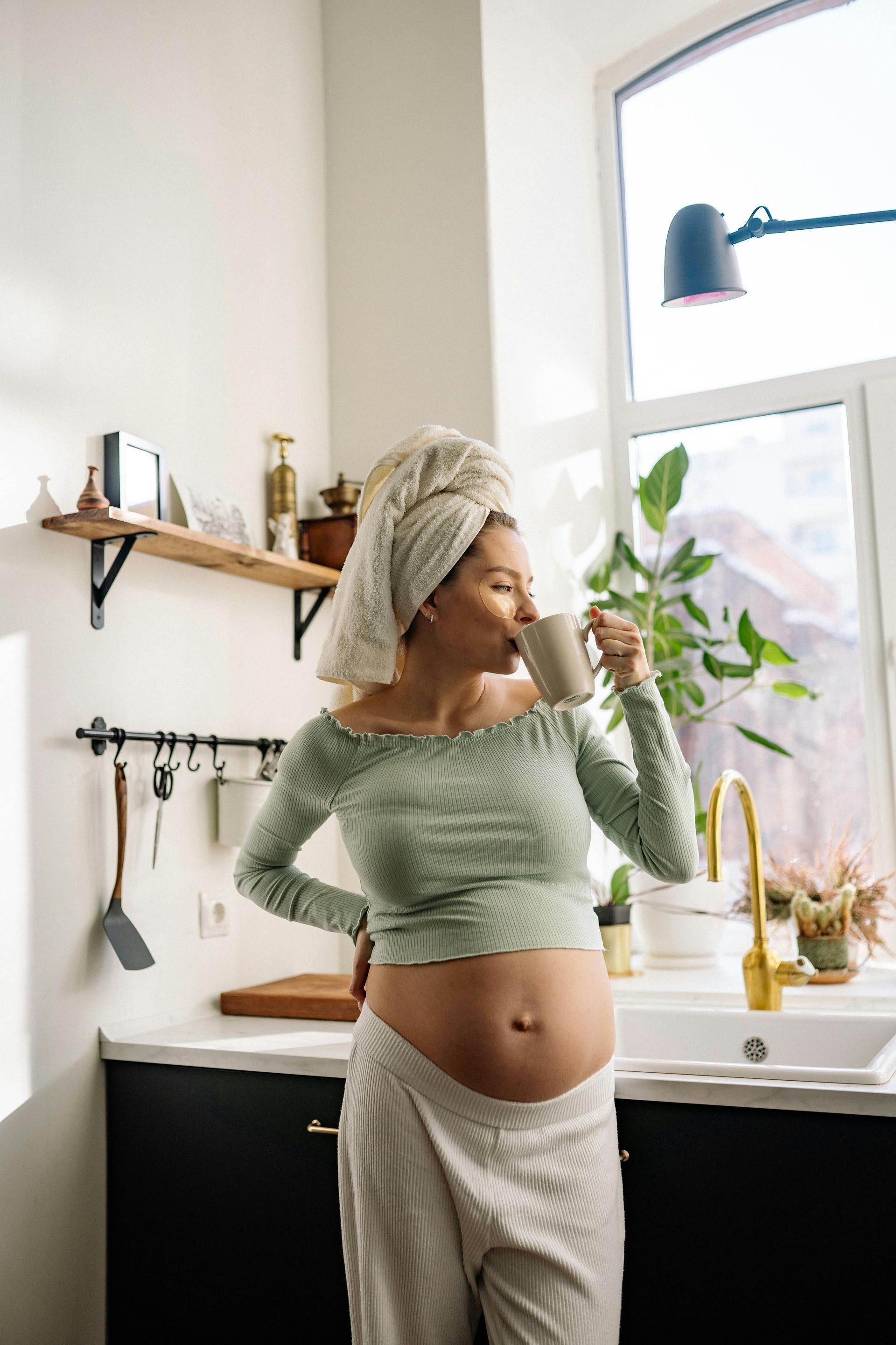 Pregnant person in kitchen, wrapped in towel, sipping from mug.