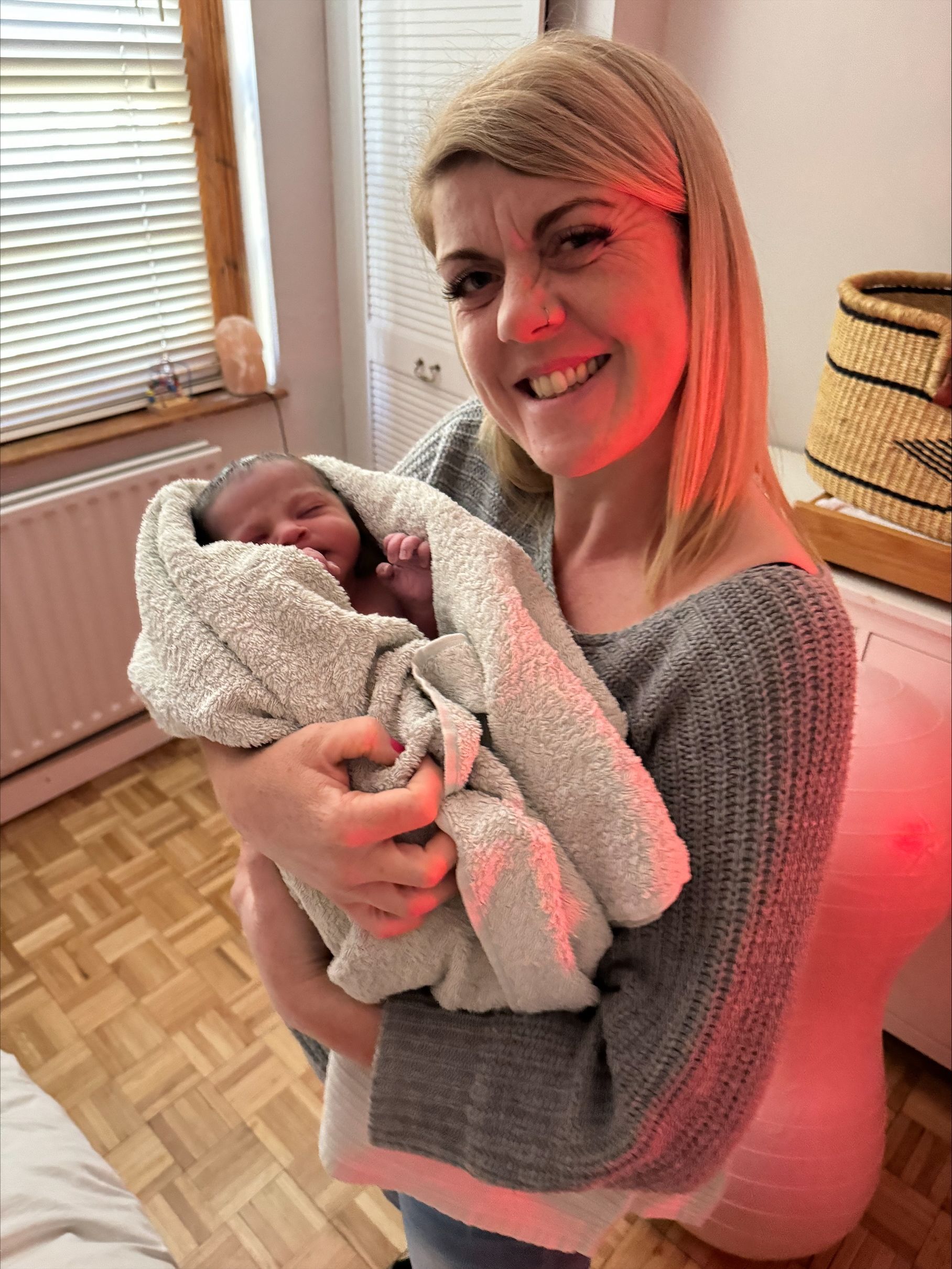 Woman smiles, holding newborn swaddled in blanket indoors. Soft red light visible.