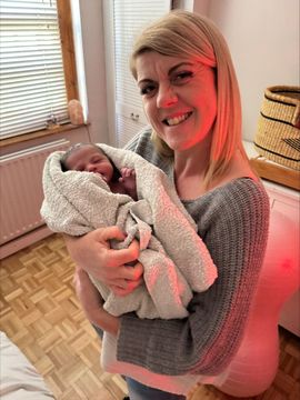 Woman holding a swaddled newborn, smiling. Indoor room, neutral colors, red light.