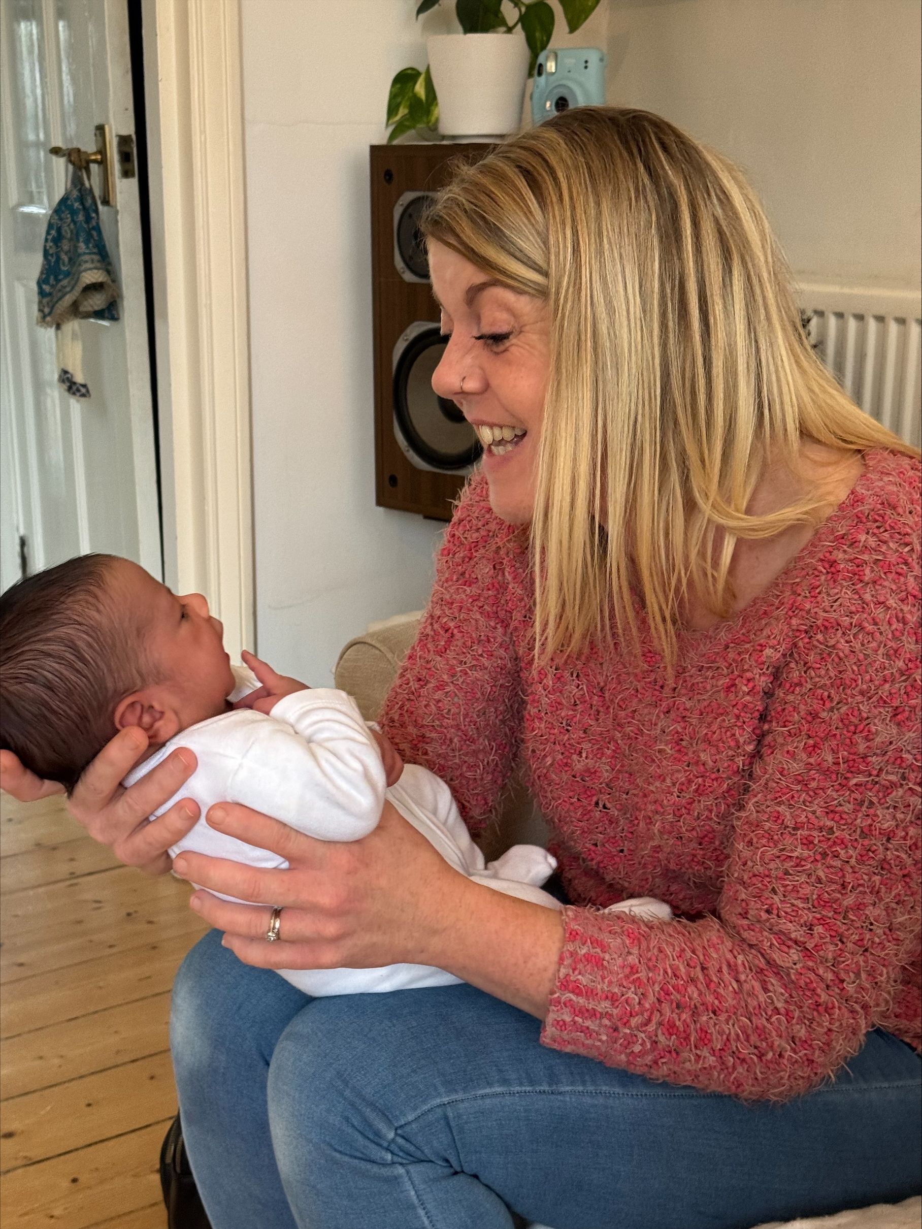 Woman smiling, holding a newborn baby in her lap. Both are indoors, baby in white onesie.
