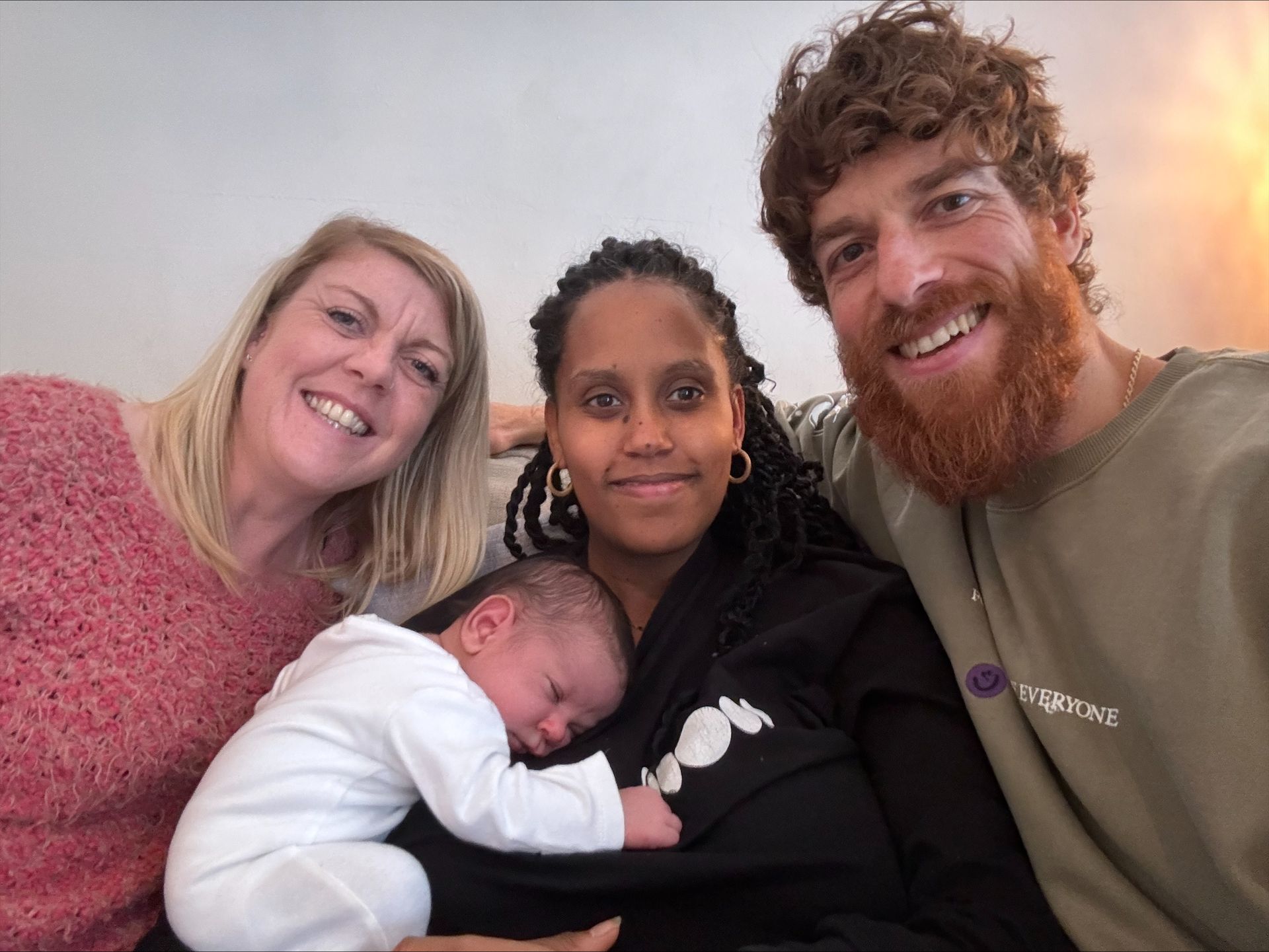 Four people smiling: a woman with a baby, and three adults posing close together indoors.