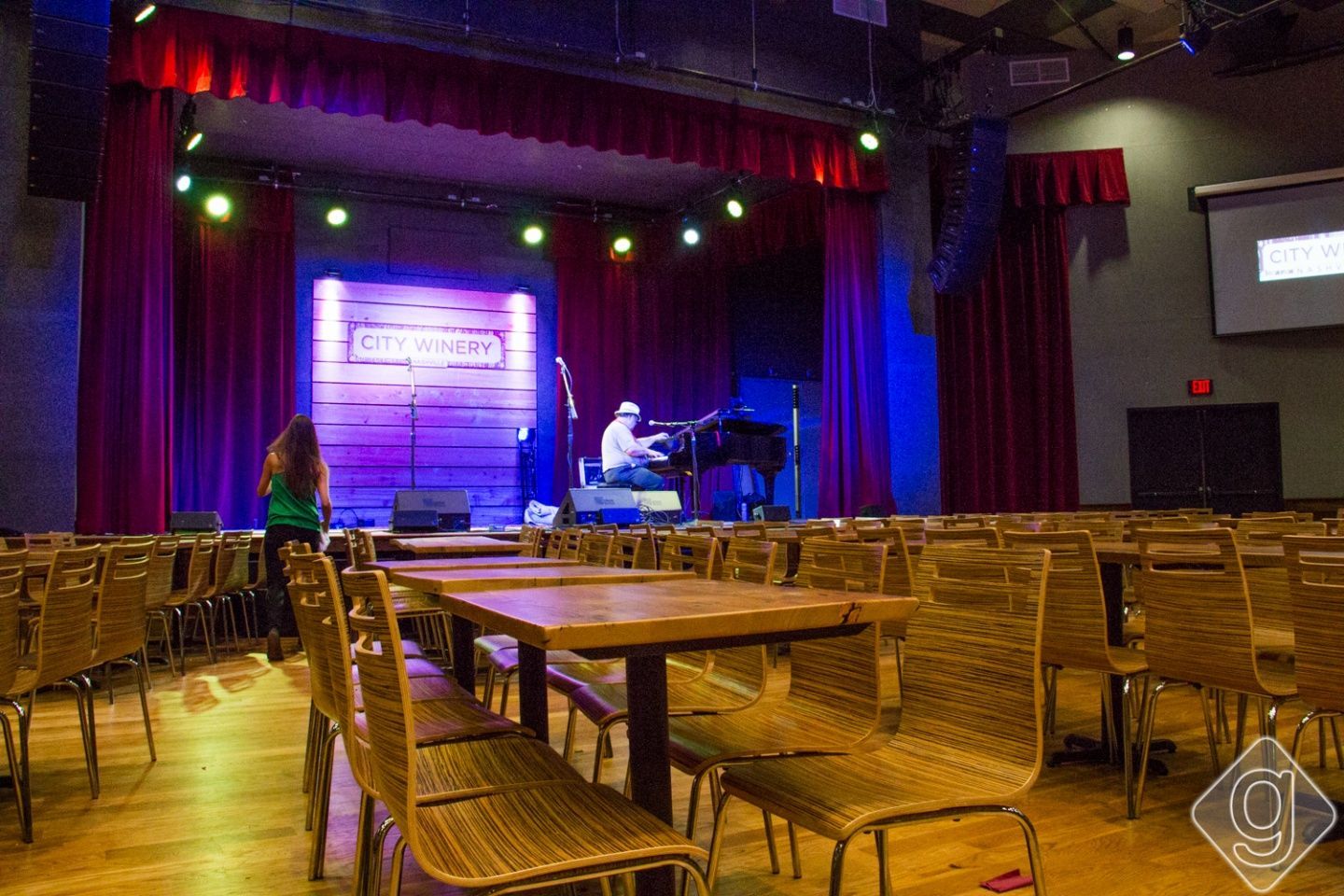 Dimly lit stage with piano and microphone, rows of wicker chairs in an empty auditorium
