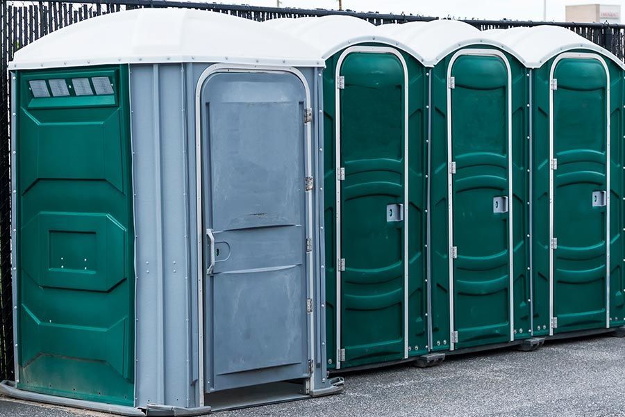 Row of green portable toilets with a white roof on gravel, one gray door in front