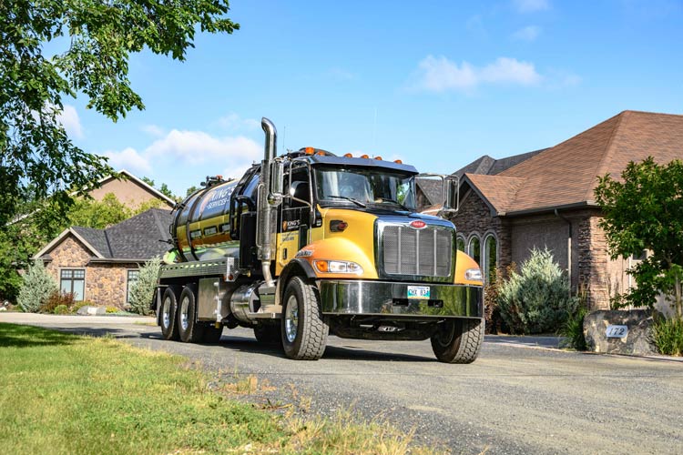 Yellow septic truck driving on a residential road. Houses and trees in the background, sunny day.