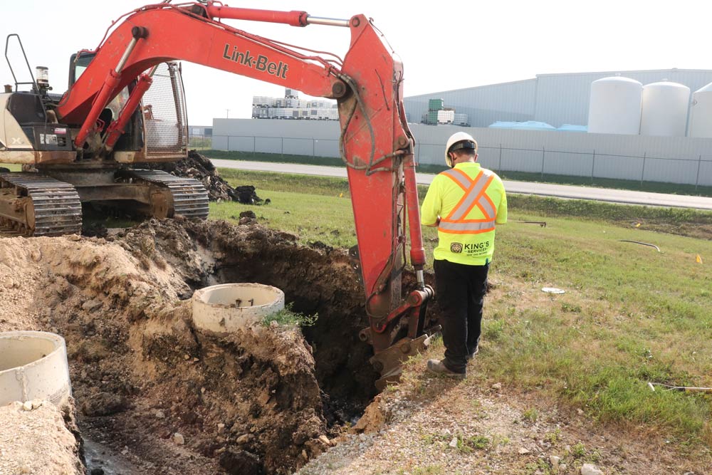 Construction worker in neon vest observes excavator digging near concrete rings outdoors.