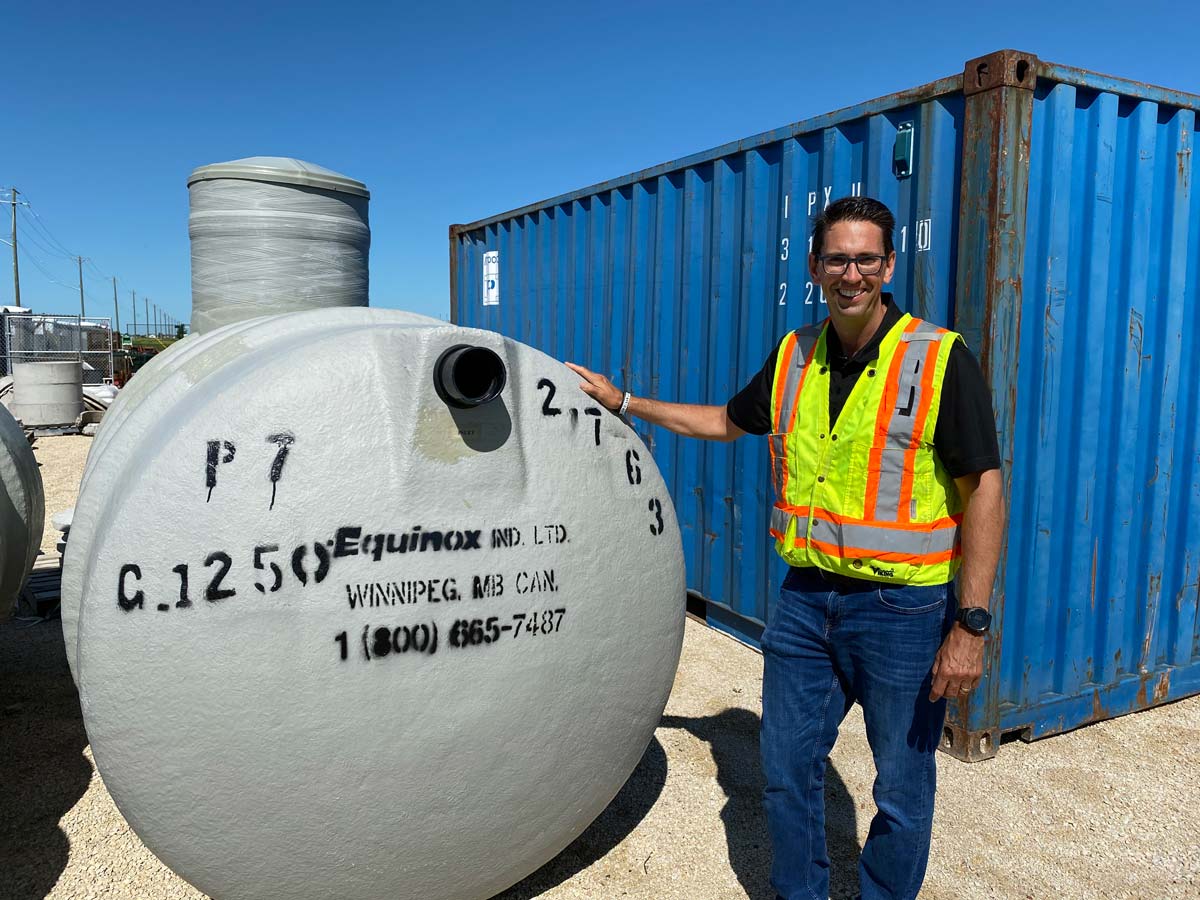 Man in safety vest stands next to a large, round tank. Blue shipping container and other tanks are in the background. Sunny day.