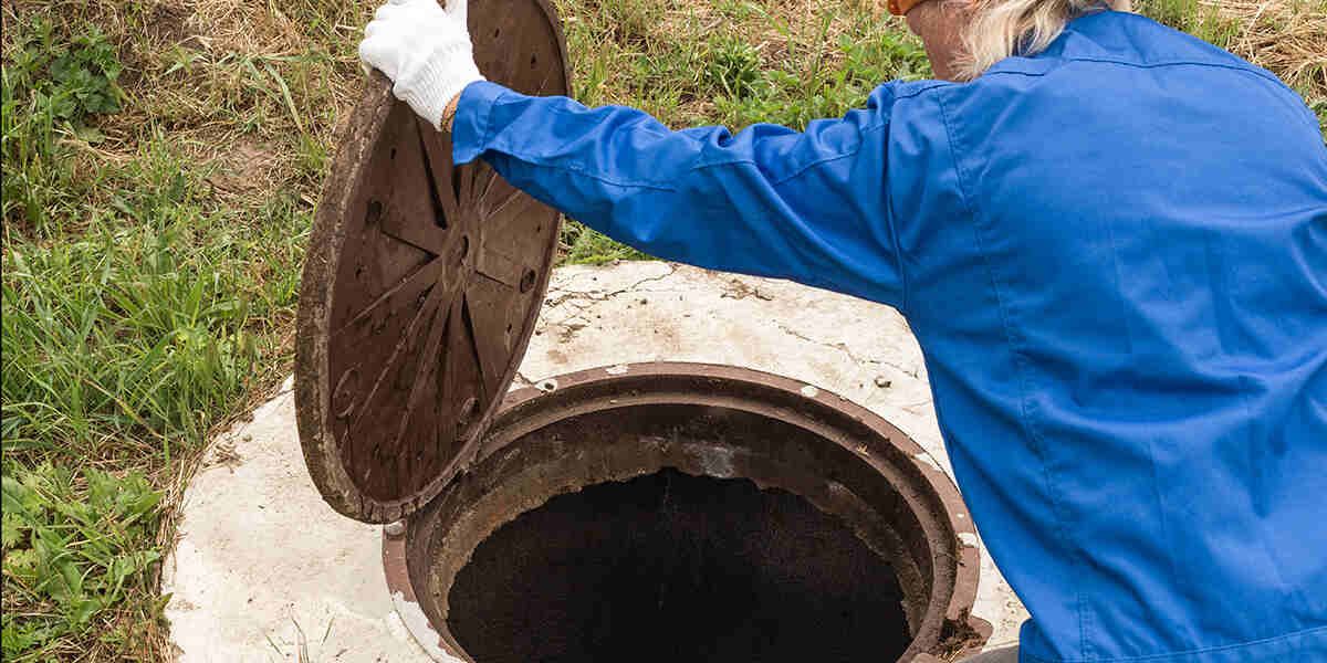 Person in blue coveralls opens a manhole cover outdoors, revealing a dark hole.