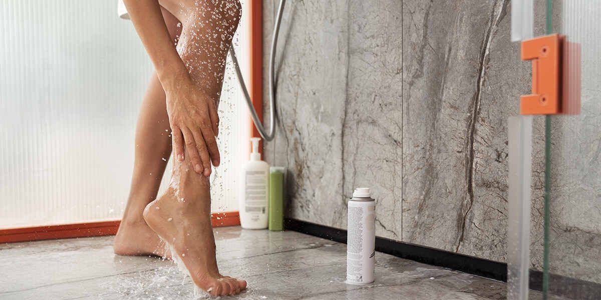 Person washes legs in a shower, water streaming down, hand touching skin. Concrete wall in background.