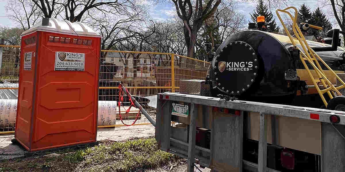 Orange portable toilet next to a trailer with a black tank, trees and construction in the background.