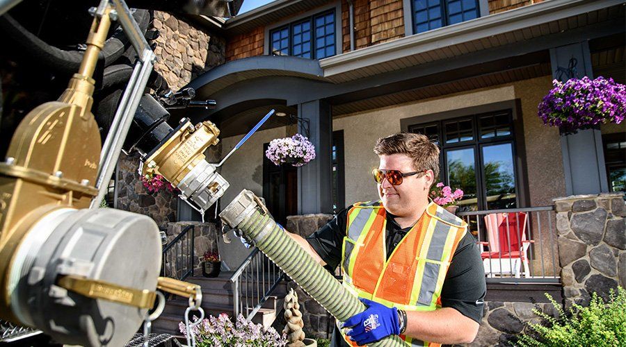 Fuel truck worker connecting hose to a house tank; orange vest, protective eyewear, sunny day.