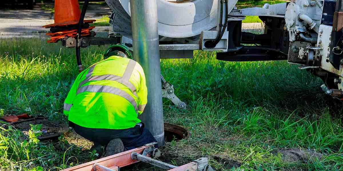 Worker in high-visibility vest kneels on grass, working on a metal pole next to a utility truck.