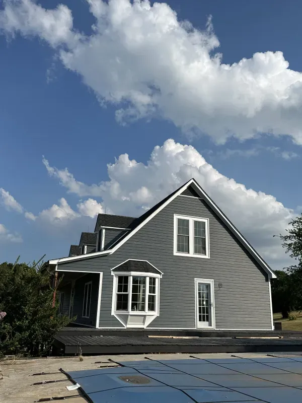 Gray house with white trim, dormers, and a bay window against a blue sky with fluffy clouds.