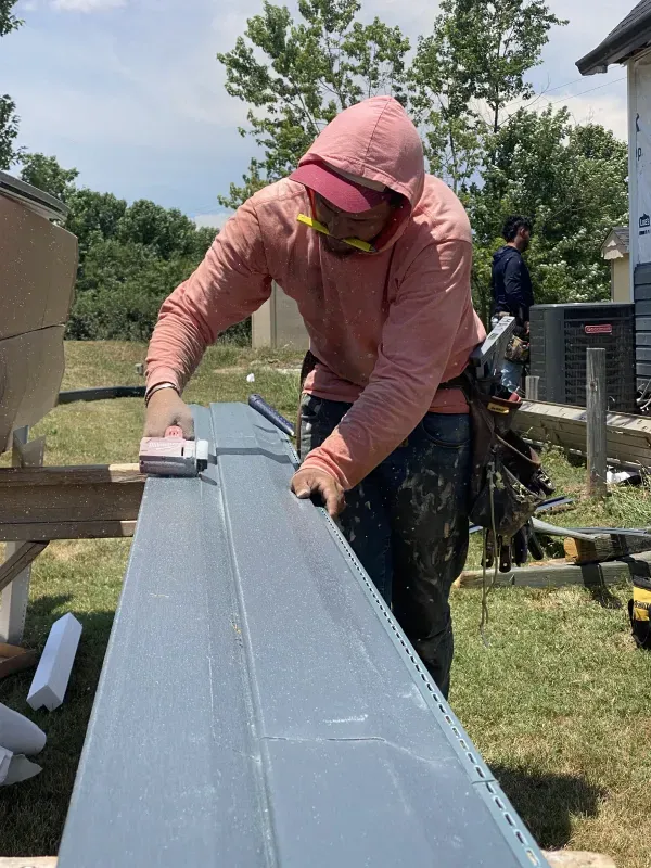 Construction worker sanding a gray siding board outdoors, wearing a pink hoodie and safety glasses.