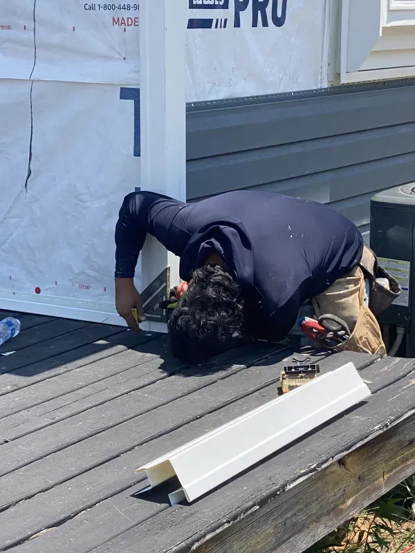 Construction worker installs siding on a structure. He's kneeling on a wooden deck. The wall has gray siding and white trim.