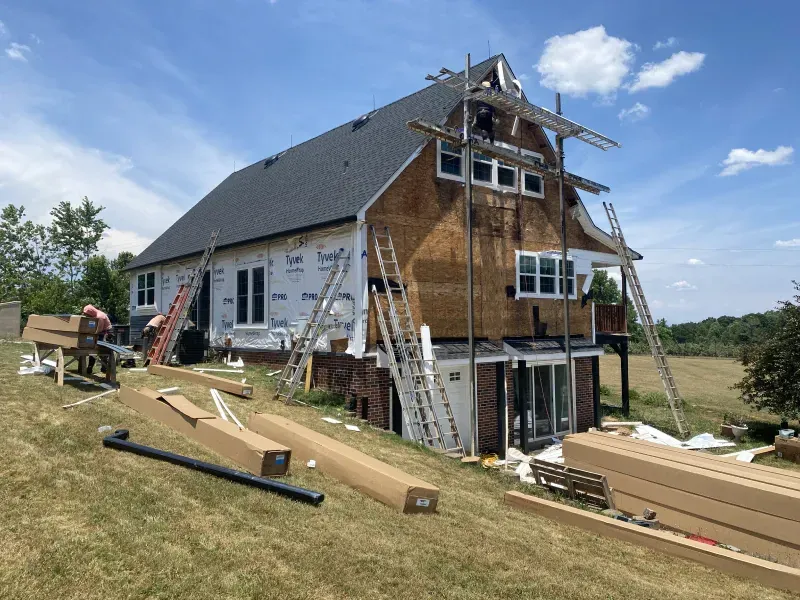 House under construction; workers on scaffolding, brown siding, blue sky, and materials on the ground.