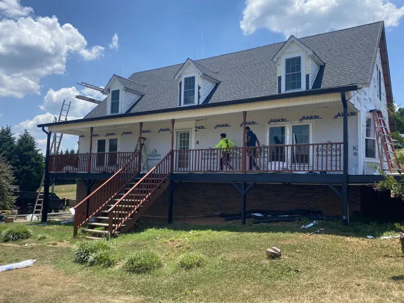 House under construction with white siding, black roof, and porch with workers.