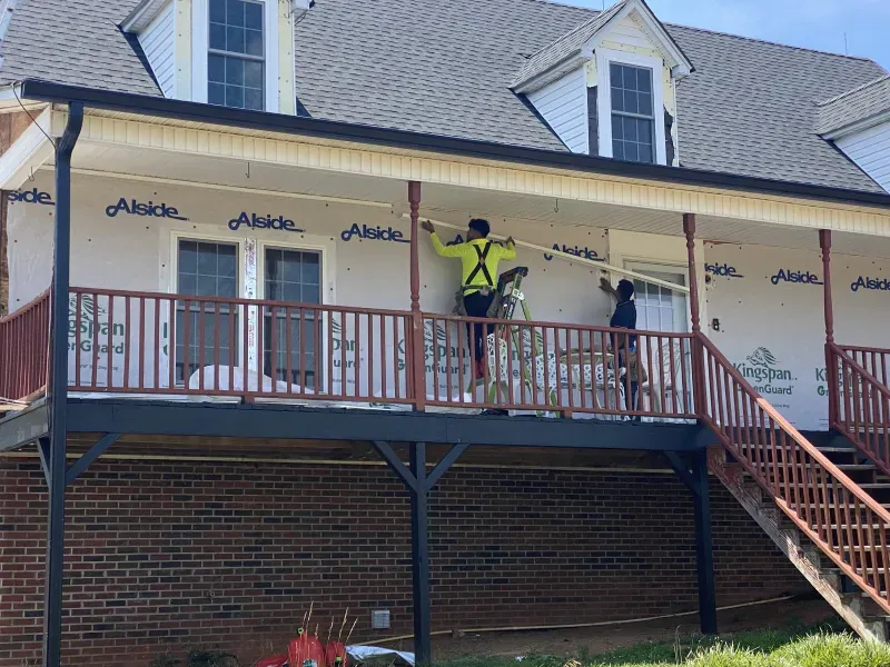 Two construction workers installing siding on a two-story building's porch with a red railing.