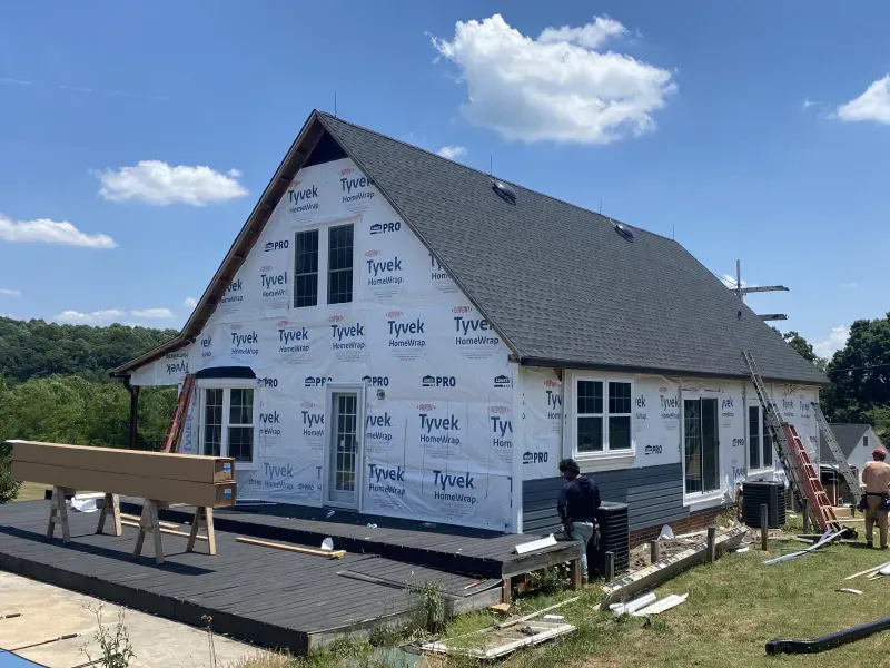 House under construction, exterior wrapped in Tyvek. Roof and dark blue siding installed. Workers on site.