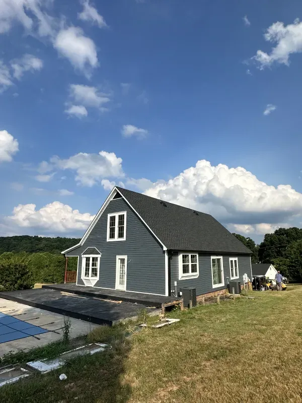 Blue-sided house with white trim under a cloudy blue sky. Includes windows, a deck, and surrounding greenery.