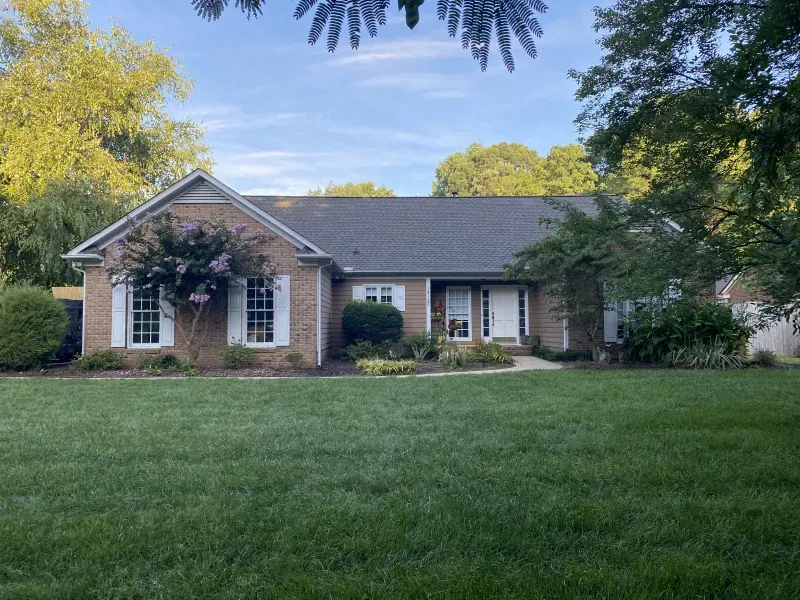 Brick house with white shutters and door, surrounded by green grass and trees.