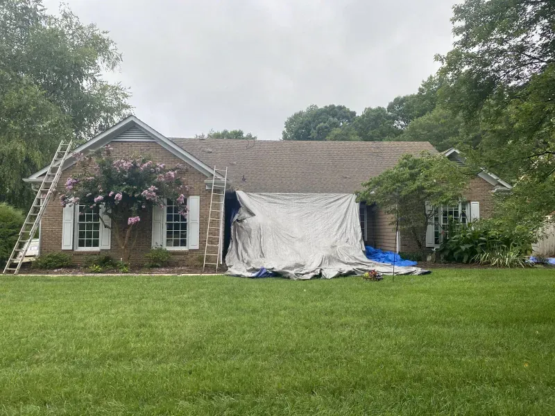 House with roof under construction, covered with a tarp. Ladders and tools are present in a green yard.