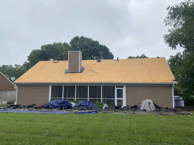 House with roof partially stripped, covered in new underlayment; tarps on ground.