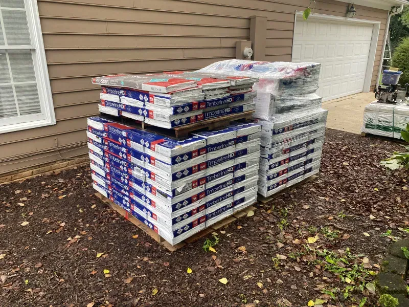Pallets of packaged roofing materials sit on a yard, next to a garage.