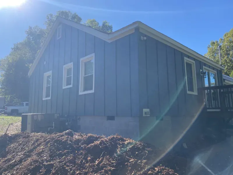 Blue-sided house with white window frames under a clear sky. A dark wood deck and mulch are in the foreground.