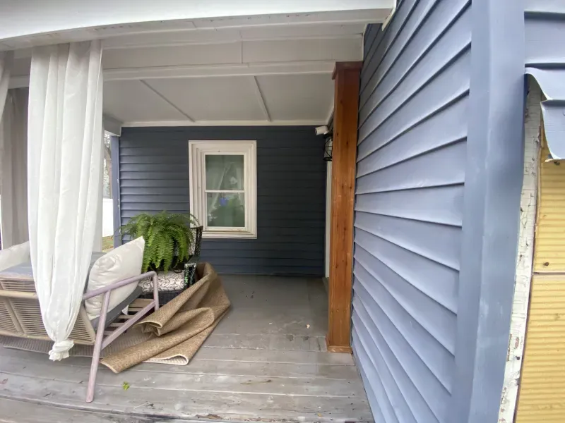 Exterior porch with blue siding, white window, and a brown column. Rug and chair present.