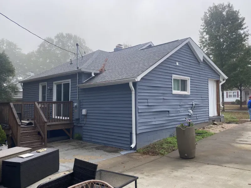 Back of a blue house with a deck, concrete patio, and overcast sky.