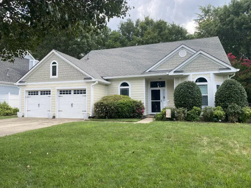 A light yellow house with a two-car garage, green lawn, and various shrubs.