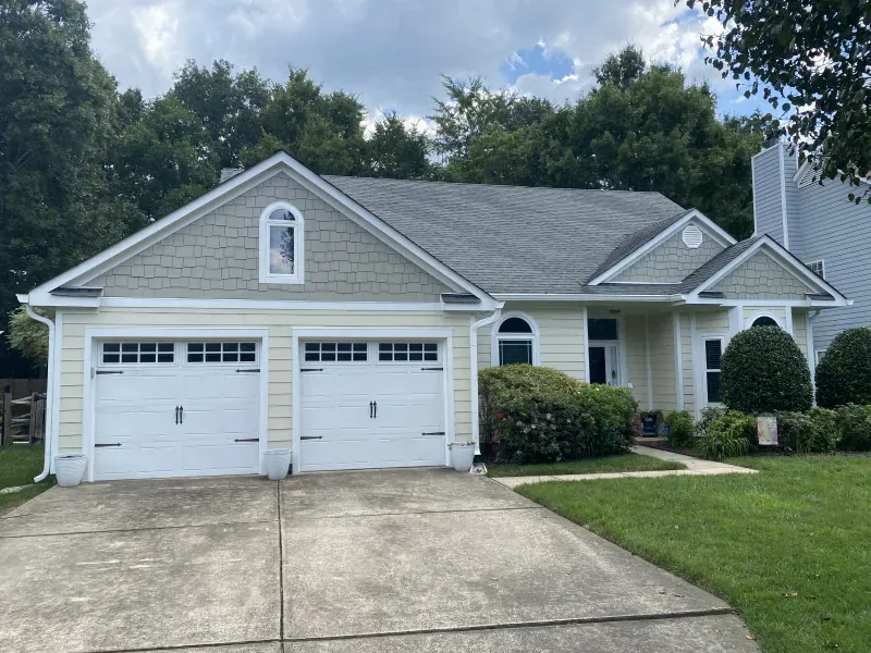 Yellow house with two-car garage, grey roof, and green front lawn. Overcast sky.