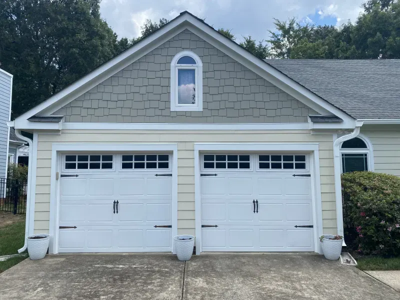 Two-car garage with white doors and beige siding, a gray shingle gable, and a small arched window.