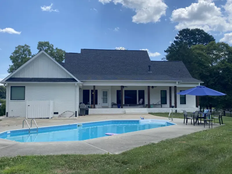 White house with pool; blue water, umbrella, patio furniture. Green lawn, trees, and a blue sky.