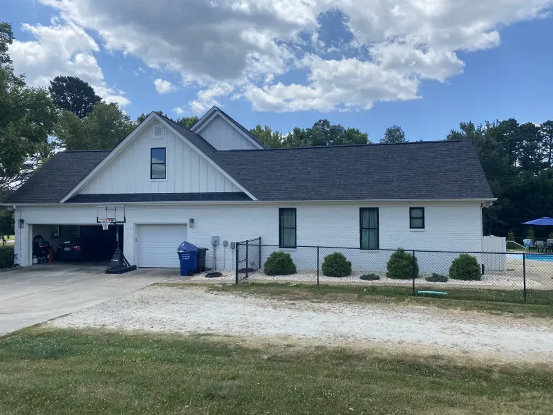 White house with black roof, attached garage, and black fence on a gravel driveway under a cloudy sky.