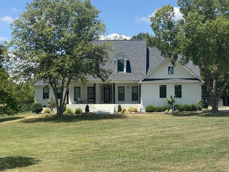 White house with a dark roof and black window frames, trees on the sides, green lawn under a blue sky.