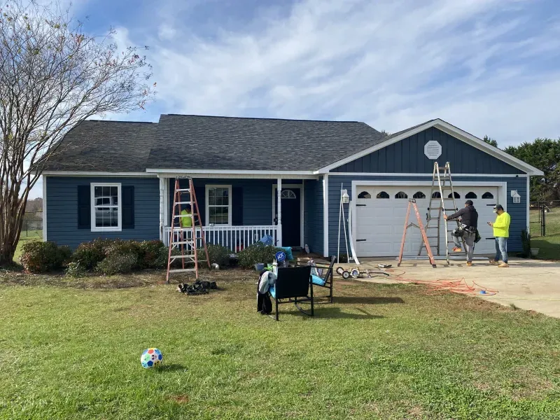 Blue house with workers on ladders; roof repair happening on a sunny day.