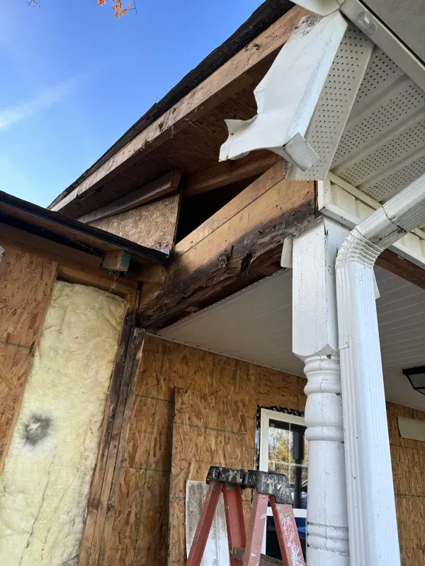 Damage to a house's roof and siding, exposing wood framing and insulation. A ladder is present.