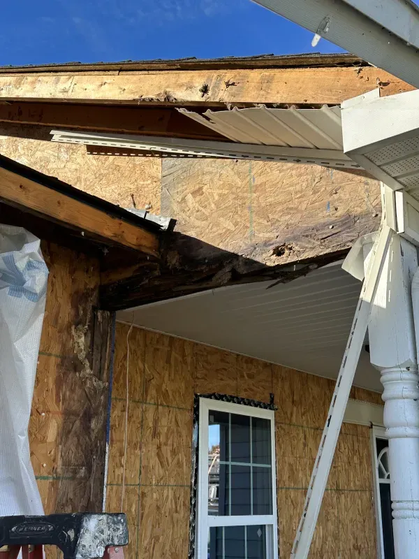 Damaged house exterior. Exposed wood framing and sheathing, damaged roof edge, missing siding and soffit.