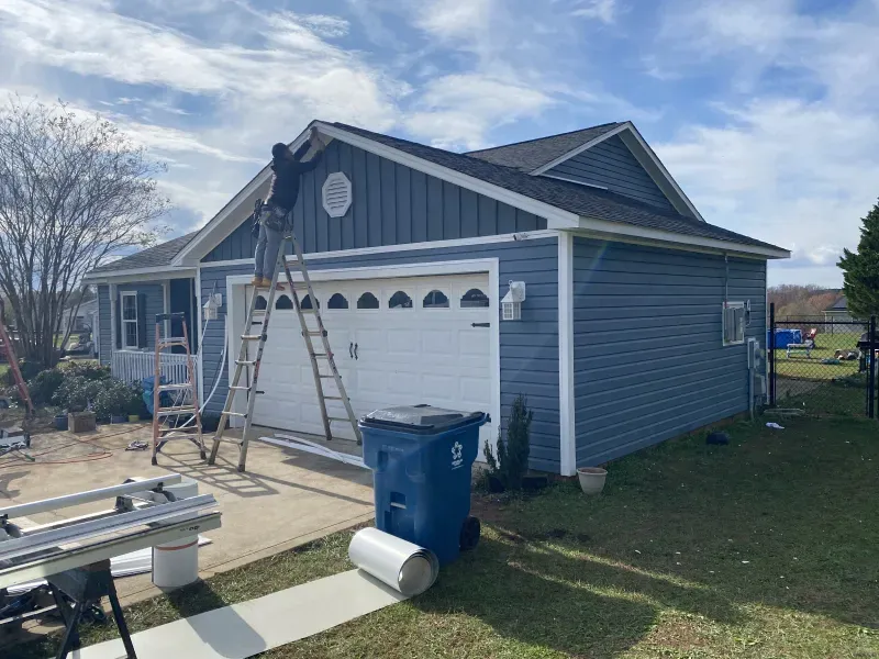 Person on a ladder working on blue siding of a house. Garage door, lawn, and blue sky in background.