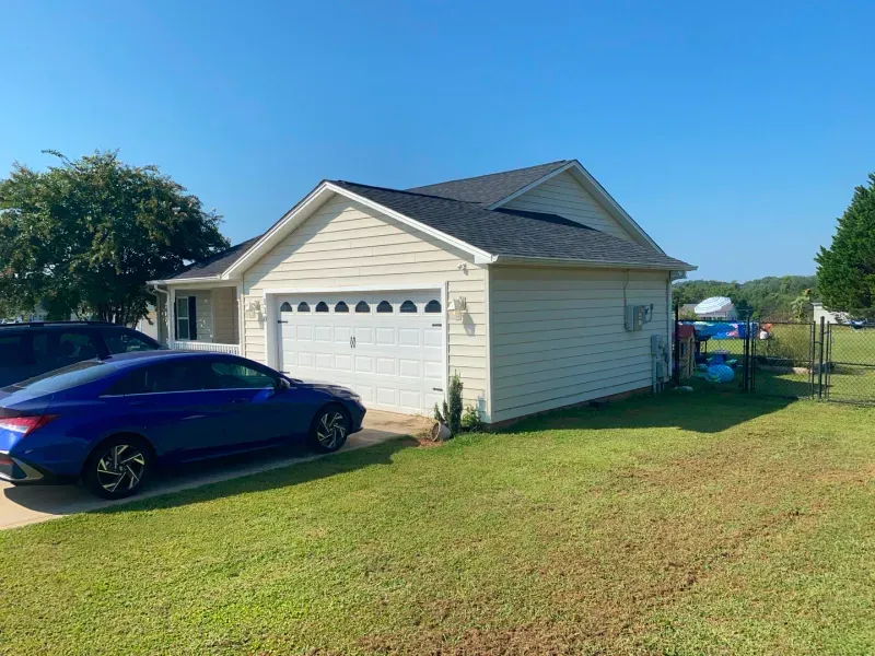 Blue car parked in front of a tan house with a white garage door, green lawn, and blue sky.