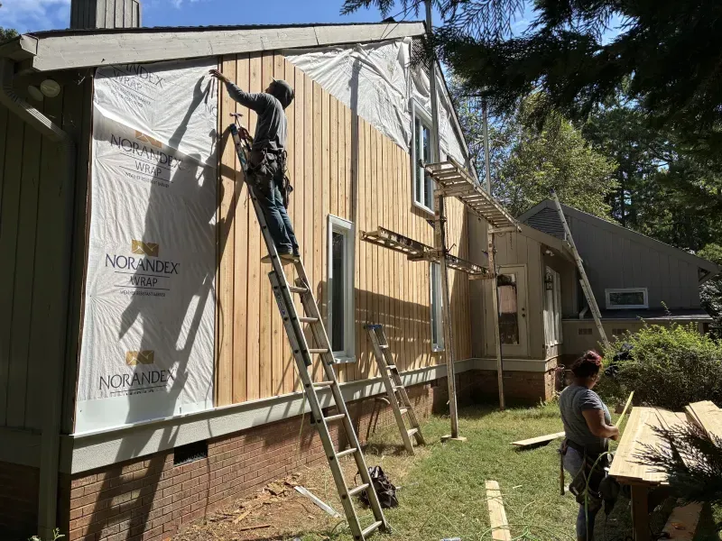 Workers installing siding on a house. One is on a ladder, the other standing near the ground.